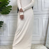 Ein langes beige Maxi-Abaya-Kleid mit langen Ärmeln und schwarzem Hijab, präsentiert vor einer weißen Paneelwand.