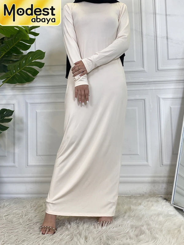 A full-length beige maxi abaya dress with long sleeves and a black hijab, displayed against a white paneled wall.