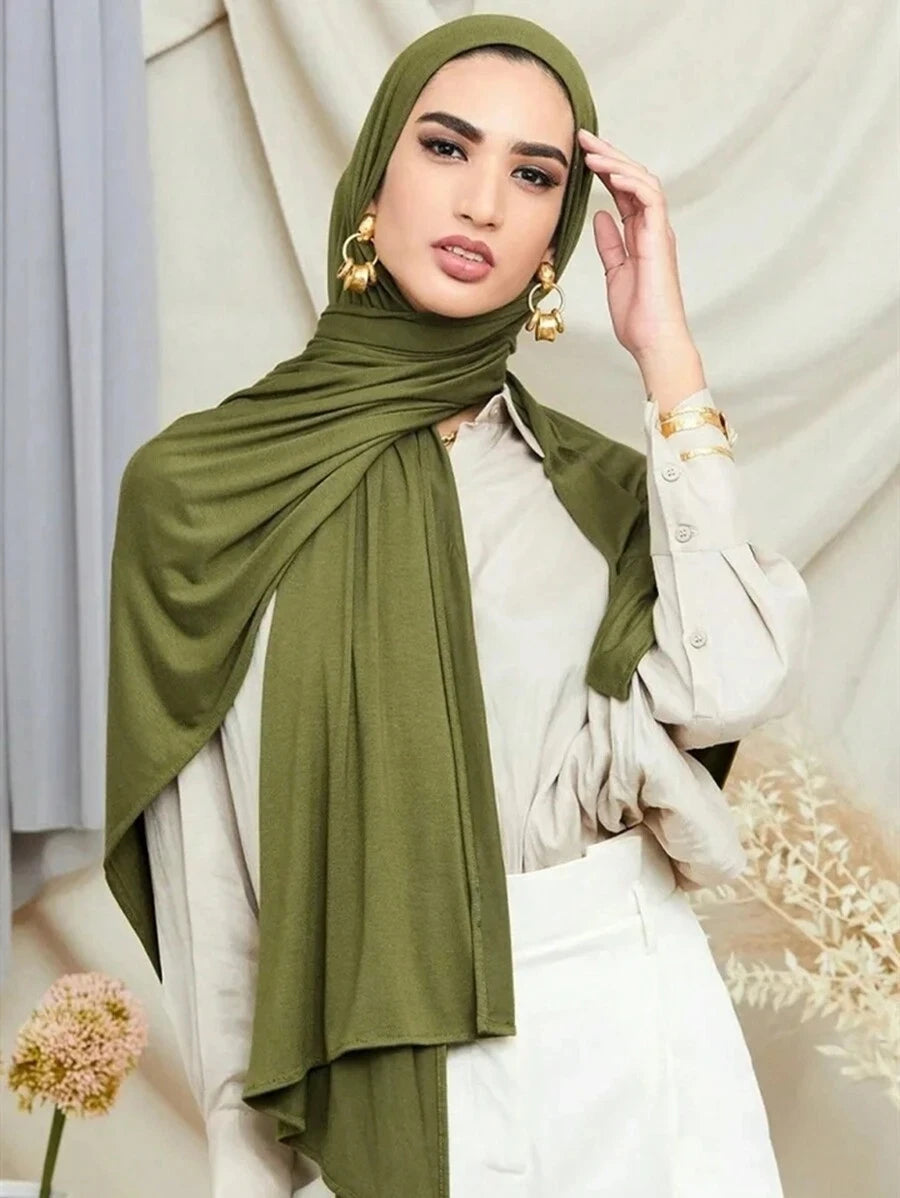 Una mujer vistiendo un pañuelo de jersey premium de modal verde oliva y una blusa blanca contra un fondo claro.