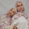 A woman wearing a pink watercolor printed scarf hijab and white top, looking directly at the camera.