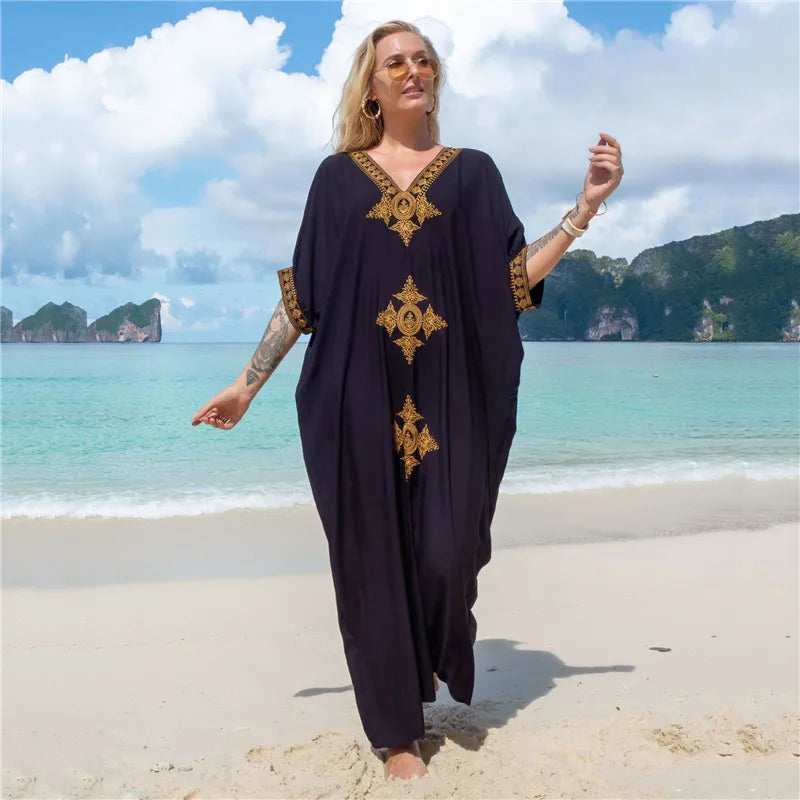 Eine Frau in einem schwarzen Kaftankleid mit goldenen Stickereien steht auf einem tropischen Strand.