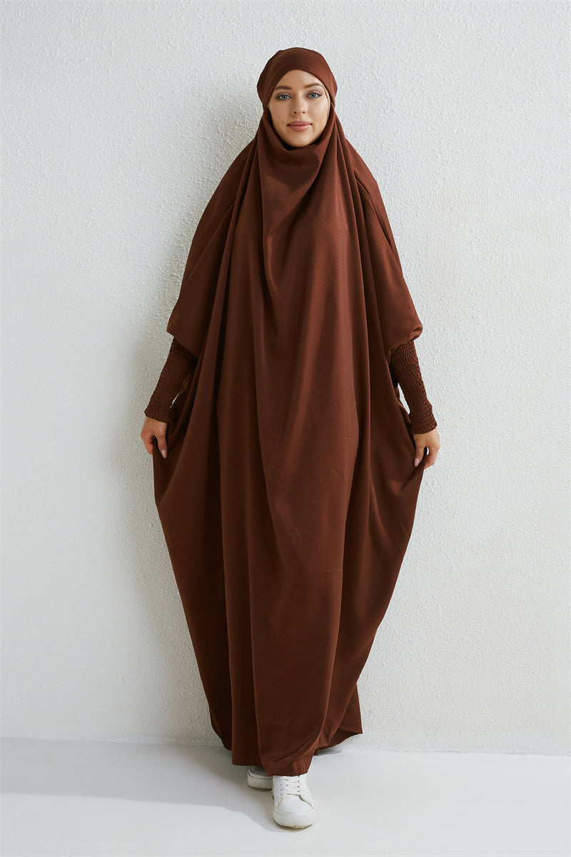 Eine Frau trägt eine braune, modische Abaya mit gerafften Ärmeln vor einer weißen Wand.