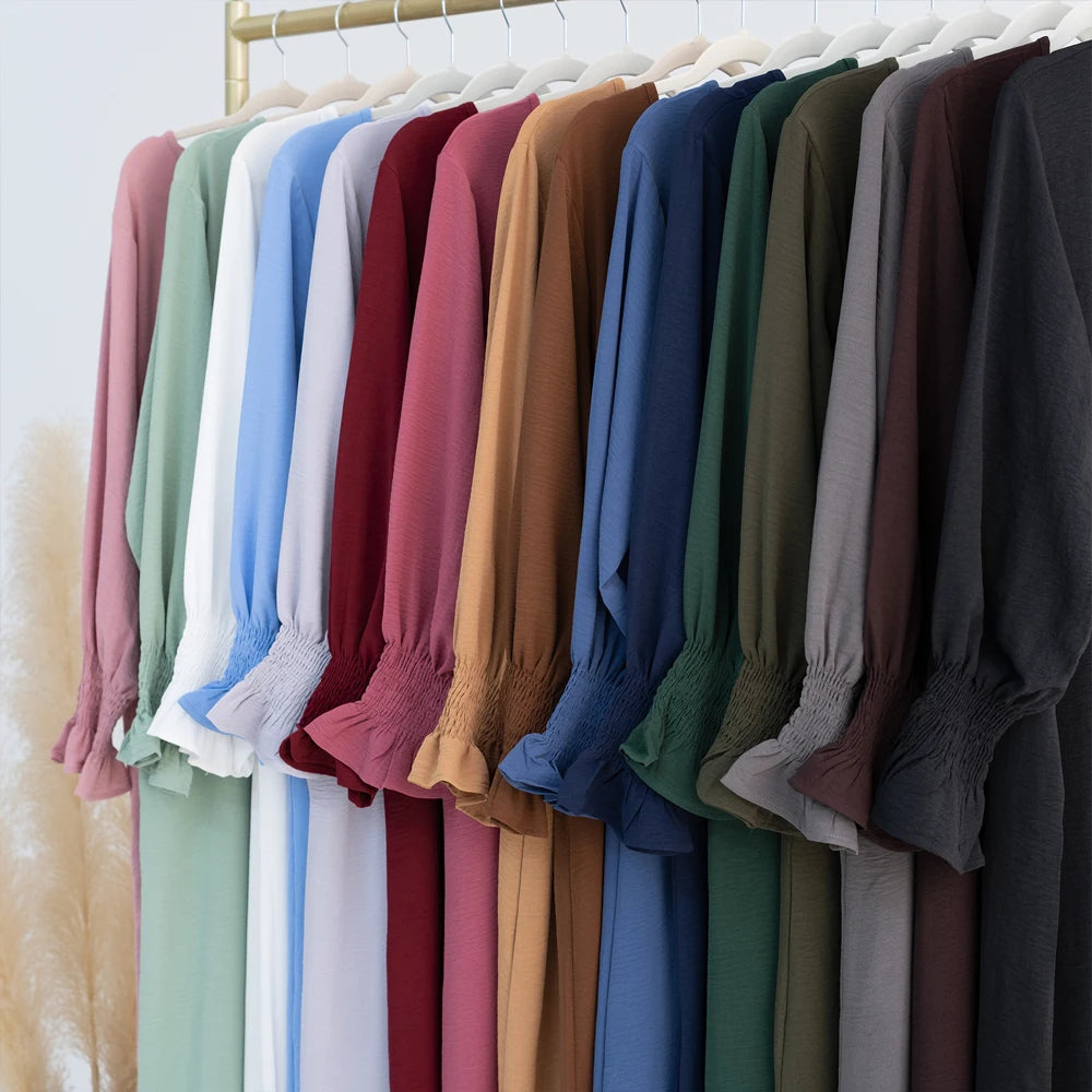 Un perchero de vestidos abaya de corte holgado en varios tonos de rosa, azul, verde y marrón colgados en perchas de madera.