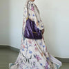 Una mujer vistiendo un caftán blanco largo y fluido con estampado floral, llevando un bolso morado.