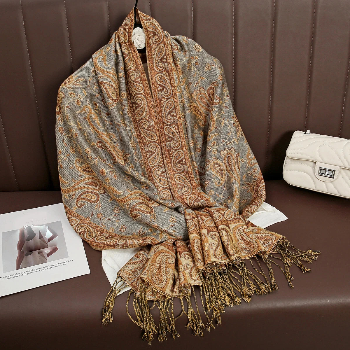 Una pashmina de lujo con patrón beige y marrón drapeada sobre una superficie de cuero marrón oscuro.