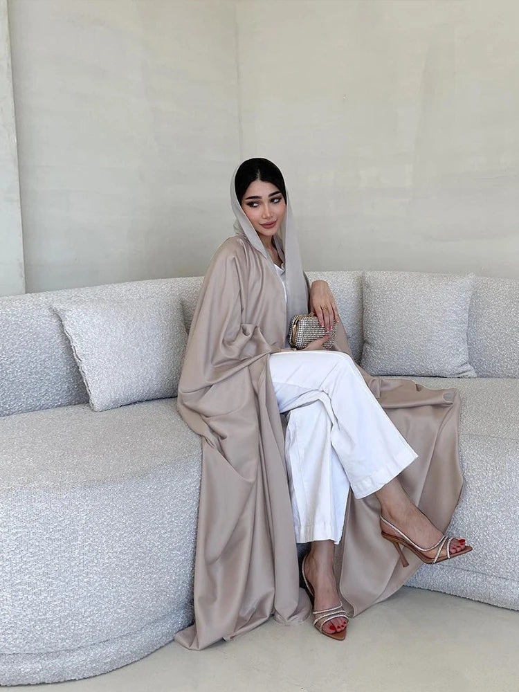 Una mujer vistiendo una abaya de satén beige con mangas de murciélago y pantalones blancos, sentada en un sofá claro.