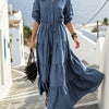 Eine Frau in einem blauen Maxi-Kleid mit Kordelzug und Rüschensaum steht auf einem Steinweg in einem sonnigen, weißgetünchten