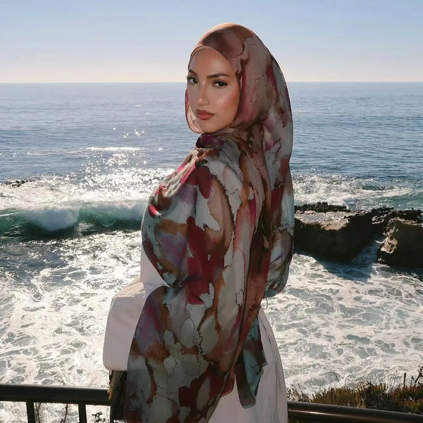 A woman wearing a watercolor chiffon instant hijab with magnetic closure stands by the ocean at sunset.
