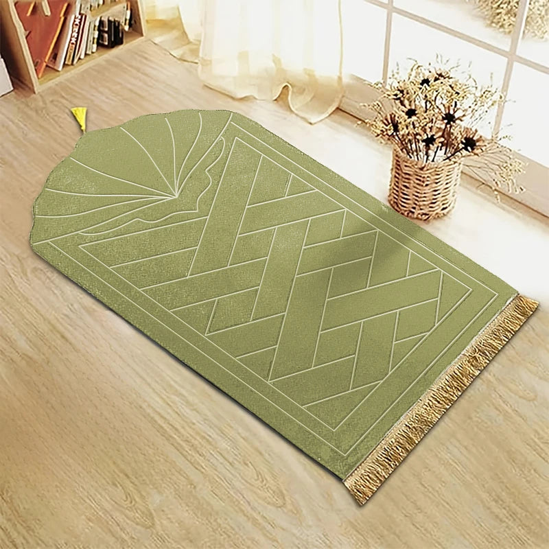 Alfombra de oración rectangular verde oliva con patrón geométrico grabado y borlas doradas sobre un suelo de madera clara.