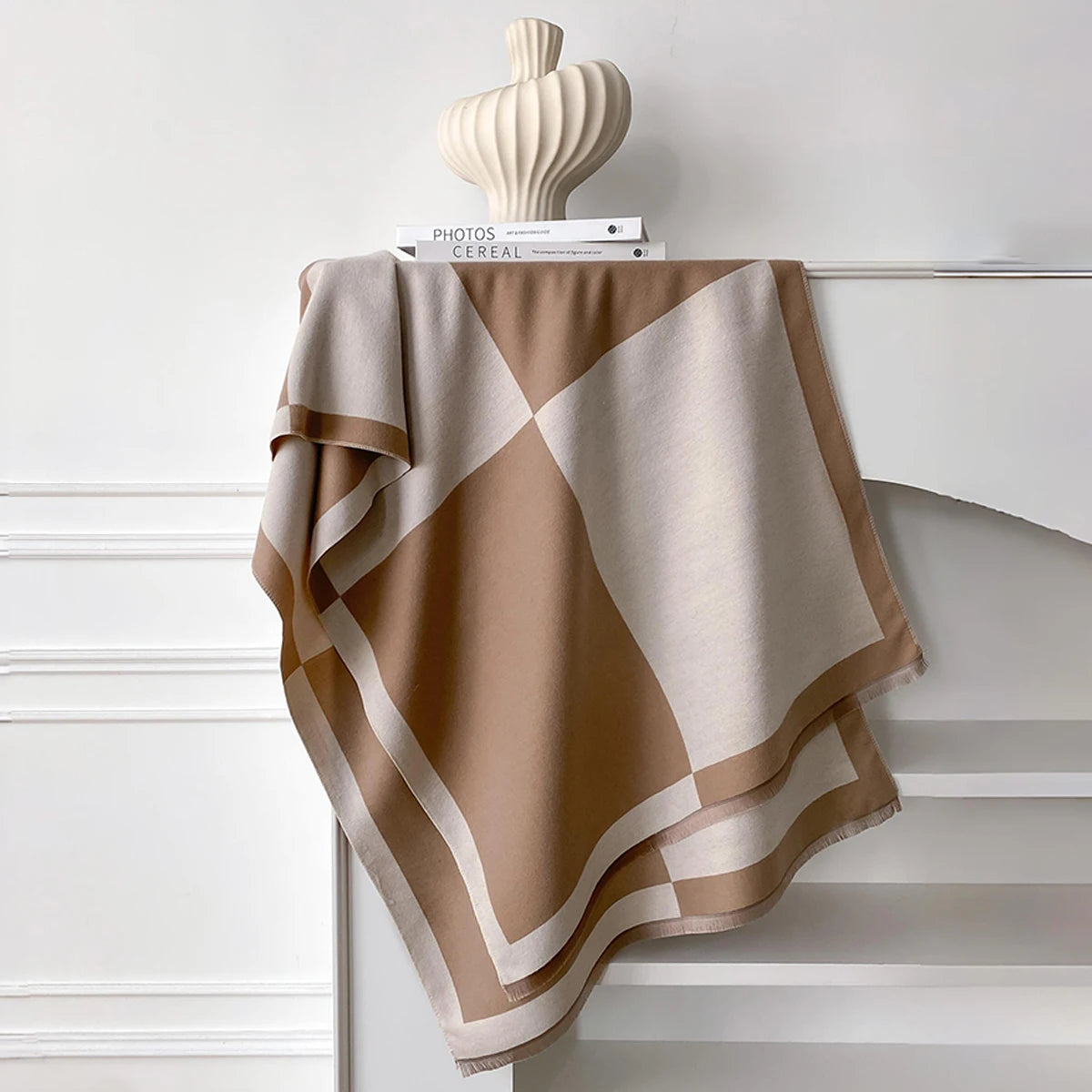 Pañuelo pashmina geométrico beige y marrón drapeado sobre una estantería blanca con un objeto de cerámica.