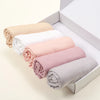 Une pile de foulards hijab unis roulés beige, blanc, rose et gris dans une boîte blanche.