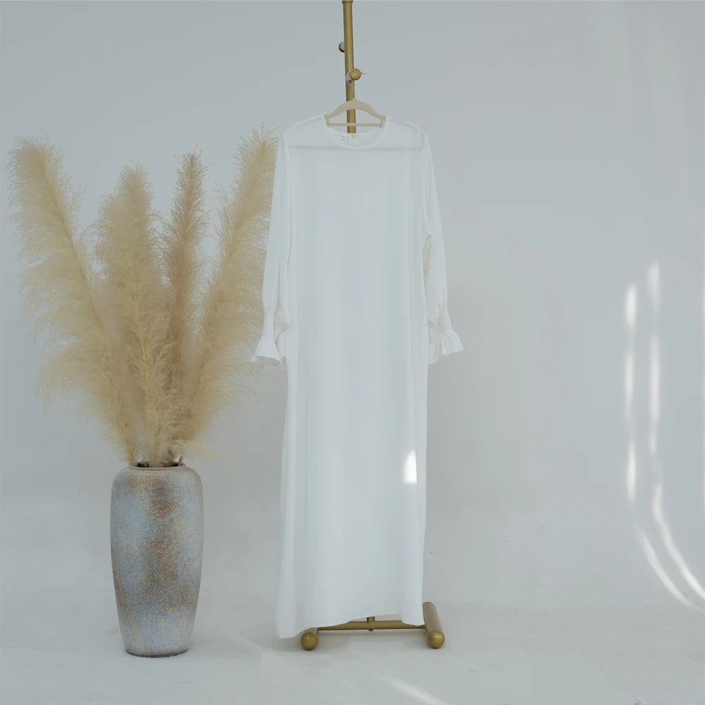 Vestido abaya blanco holgado con puños fruncidos colgado en percha de madera contra fondo gris claro.