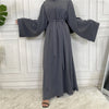 Lange, graue, elegante Abaya für Frauen mit weiten Ärmeln vor einer weißen Paneelwand.