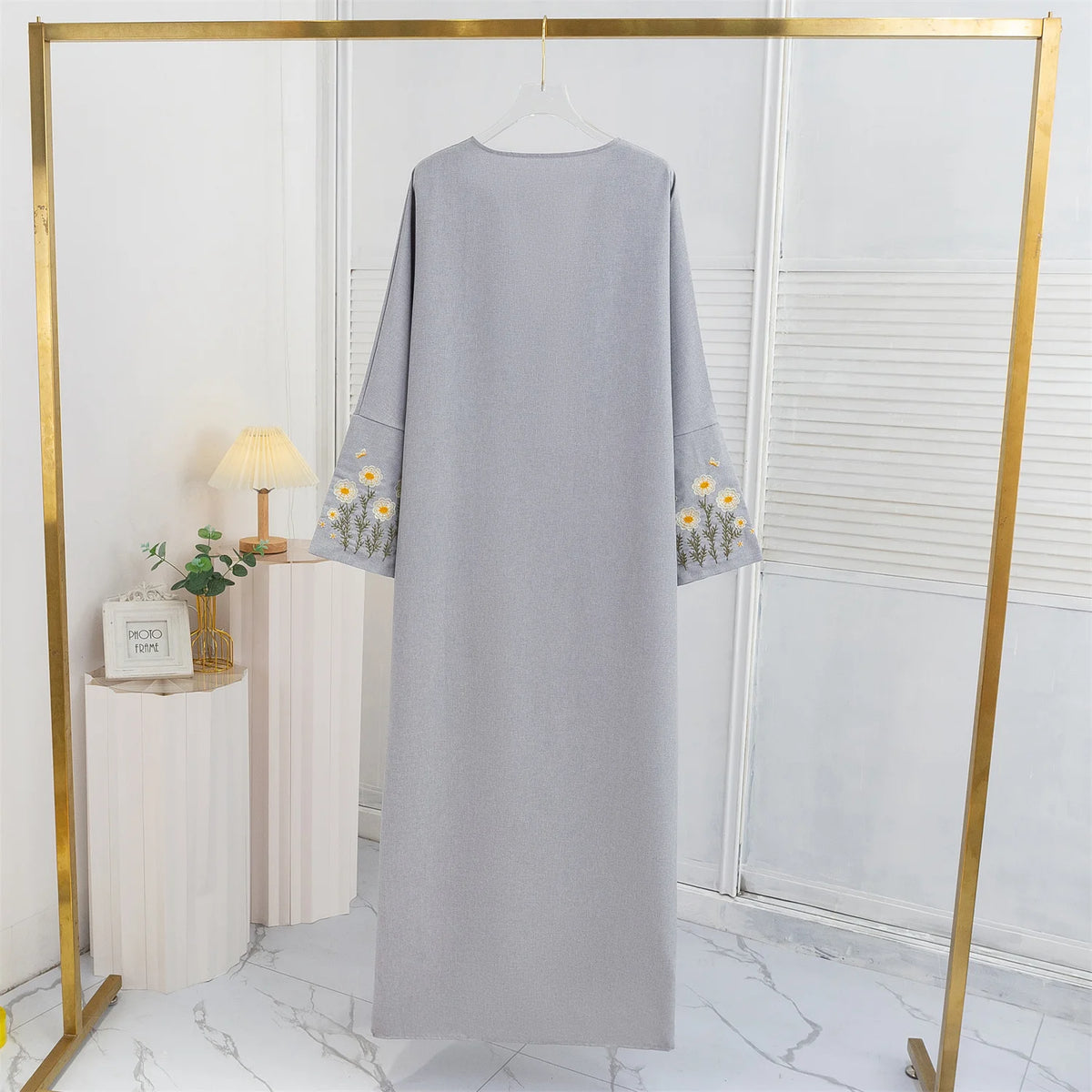 Lange, hellgraue Abaya mit Blumenstickerei auf einem goldenen Bügel in einem hellen Raum.