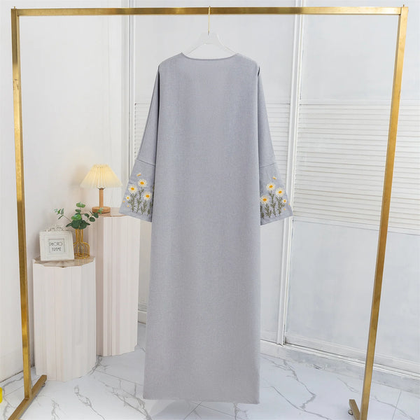 A long, light gray abaya with floral embroidery displayed on a gold hanger in a bright room.