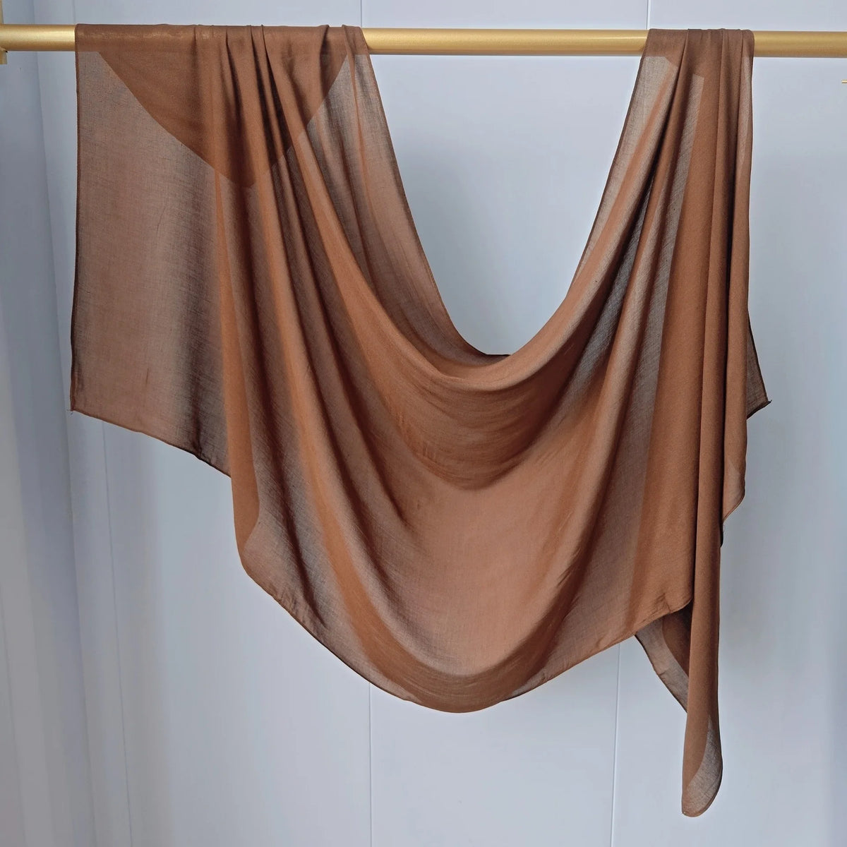 Un foulard modal brun solide drapé, suspendu sur un bâton en bois contre un mur blanc.