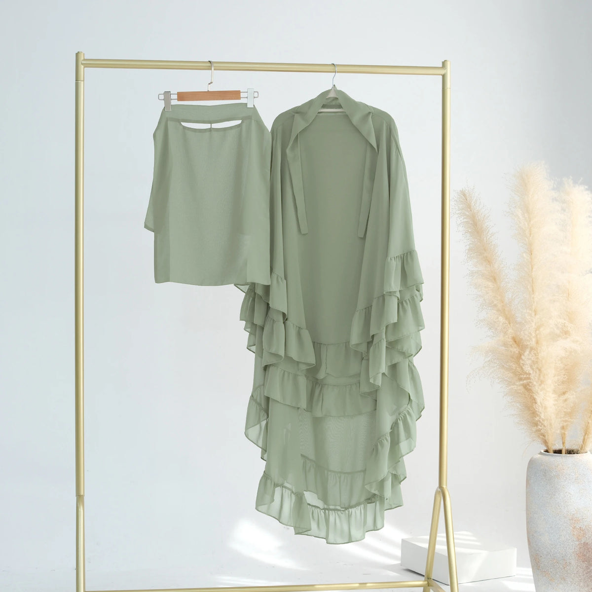 A light sage green ruffled two-layer chiffon hijab displayed on a gold hanger within a rectangular frame.