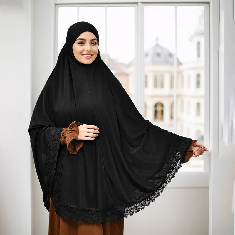 Eine Frau trägt einen schwarzen Instant-Hijab mit Spitzenrand und ein braunes Kleidungsstück, steht drinnen.