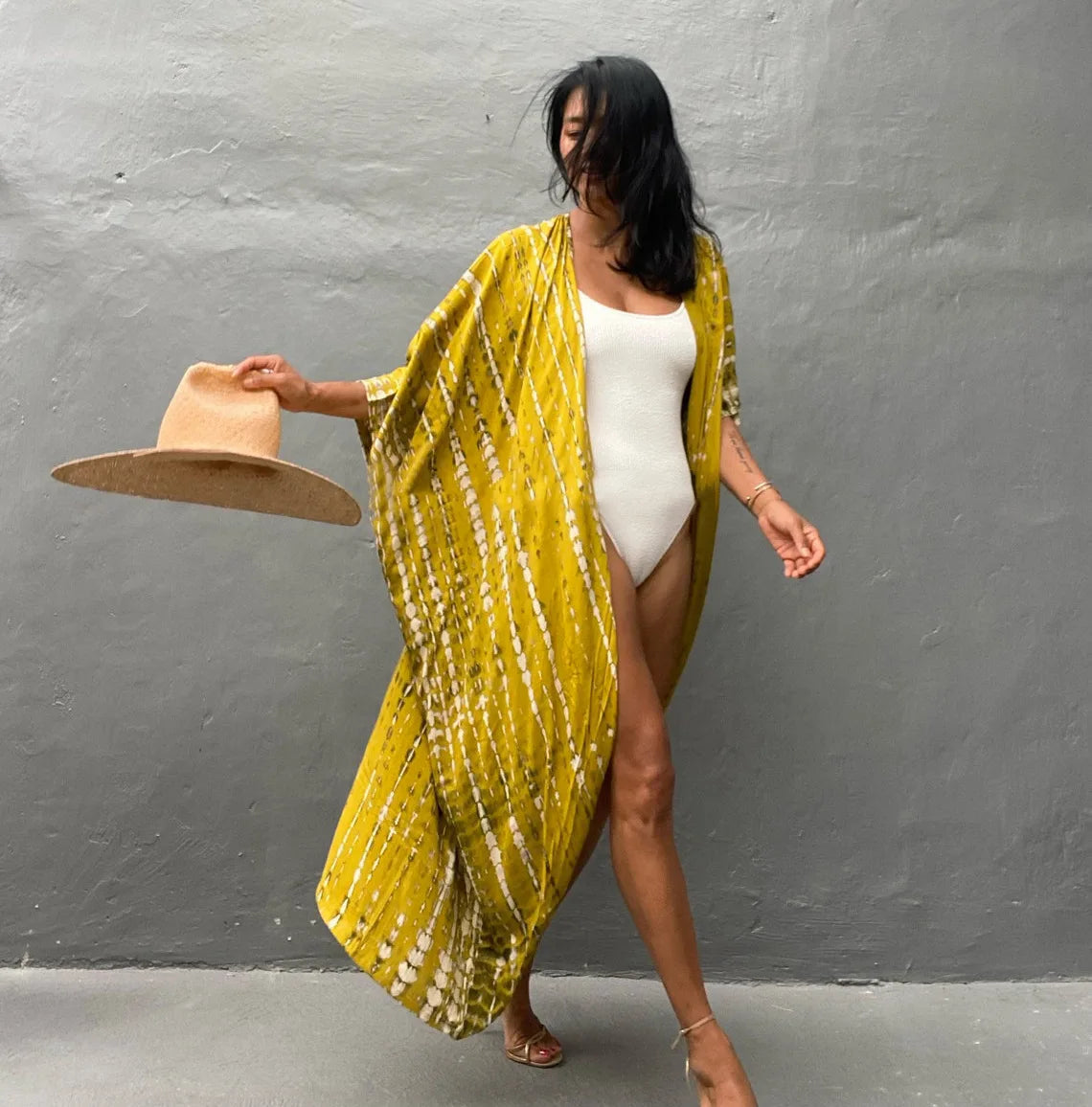 Eine Frau trägt ein gelb gemustertes Kimono-Überkleid über ein weißes Kleid und hält einen Strohhut.