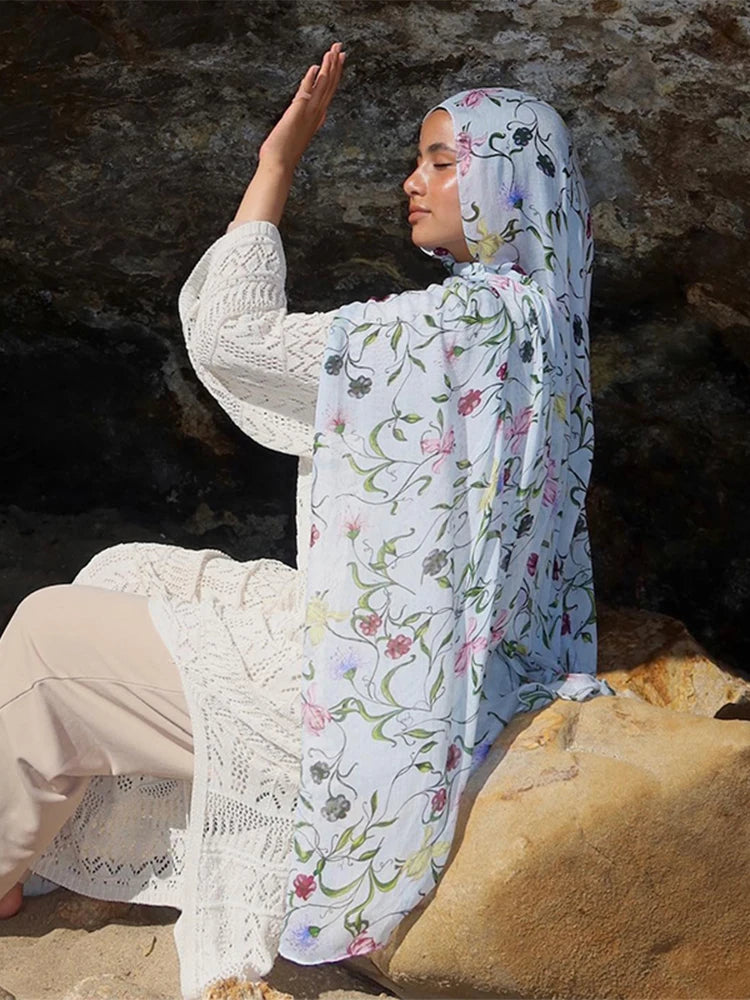 Une femme portant un hijab à imprimé aquarelle bleu clair et un haut en crochet blanc, assise sur des rochers.