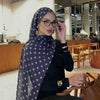 Eine Frau mit blau-weißer Polka-Dot-Stoffschal und Brille sitzt an einem Holztisch in einer modernen Innenraumumgebung.