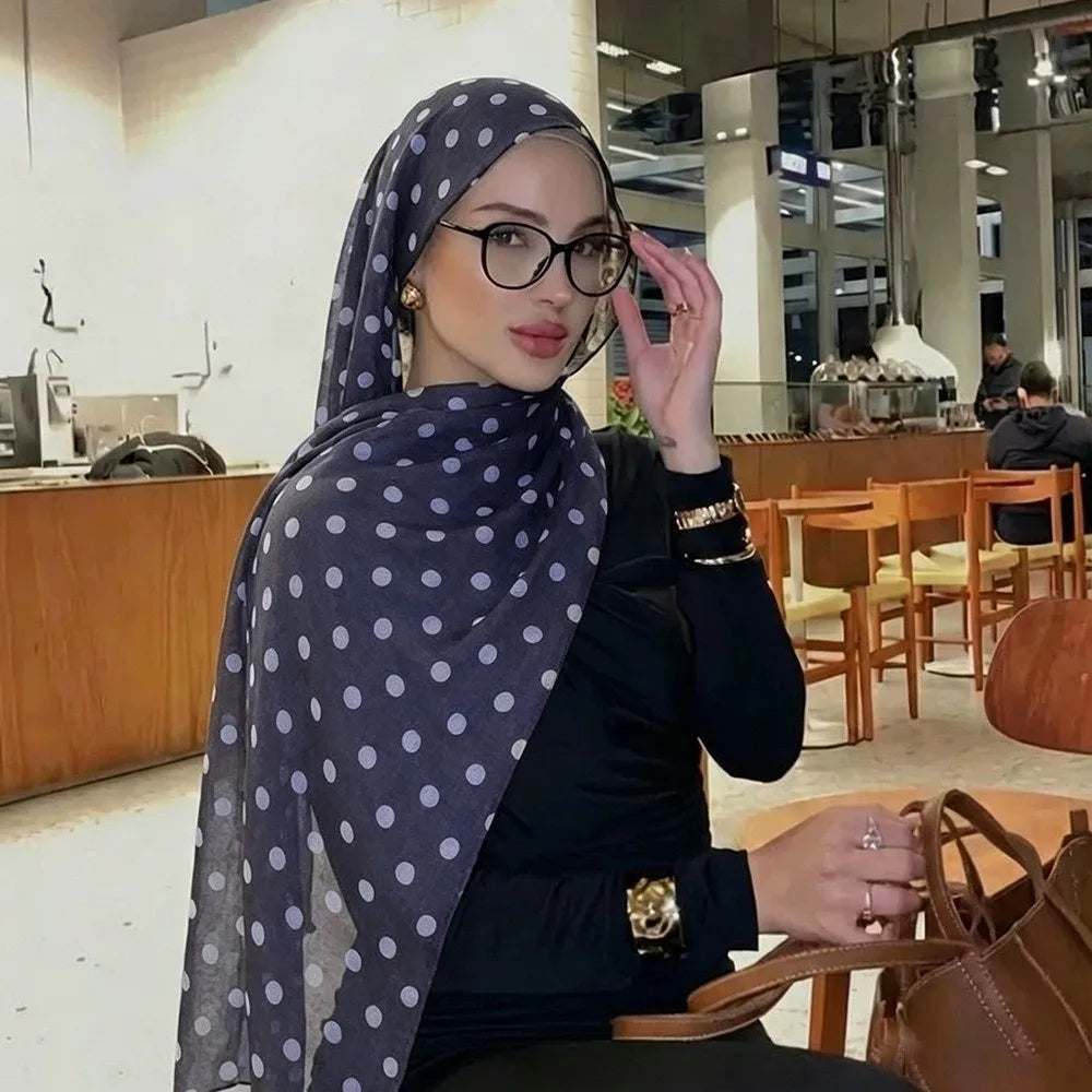 Eine Frau mit blau-weißer Polka-Dot-Stoffschal und Brille sitzt an einem Holztisch in einer modernen Innenraumumgebung.