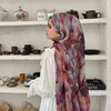 Eine Frau mit aquarelliertem Hijab-Schal steht vor weißen Regalen mit Keramik und geflochtenen Körben.