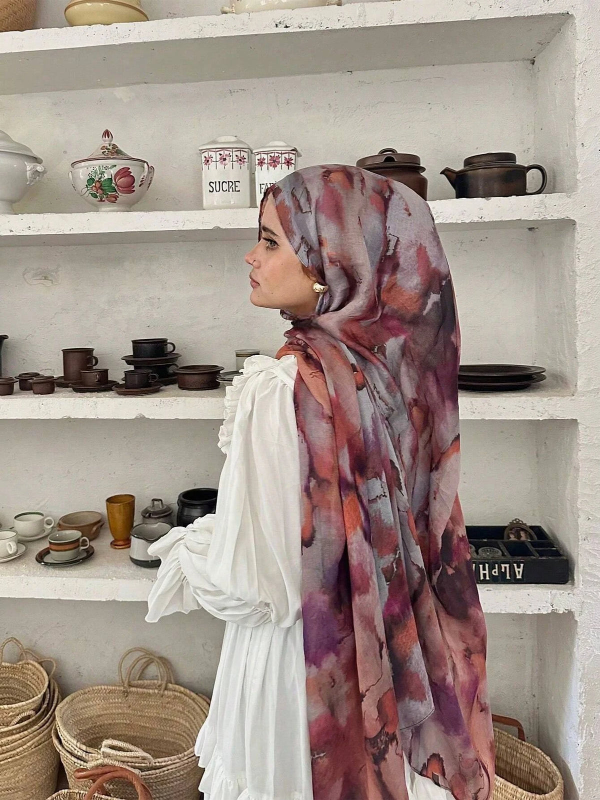 Eine Frau mit aquarelliertem Hijab-Schal steht vor weißen Regalen mit Keramik und geflochtenen Körben.