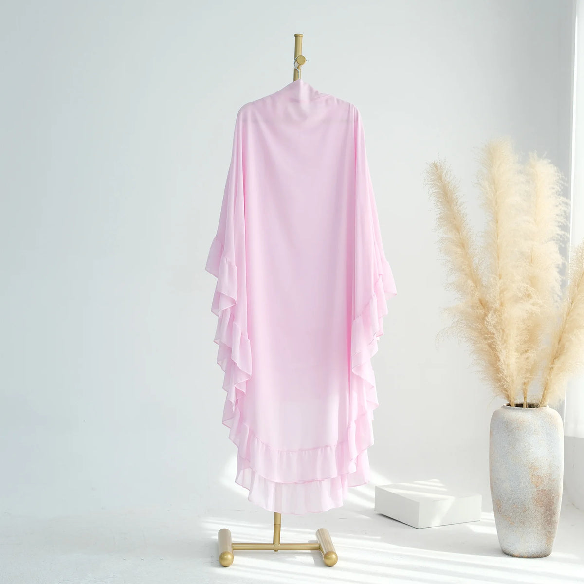 A pink ruffled two-layer chiffon hijab is displayed hanging on a wooden hanger against a white background.