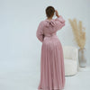 Une femme portant une abaya rose poudré fluide avec une ceinture ornée, debout sur un fond blanc.