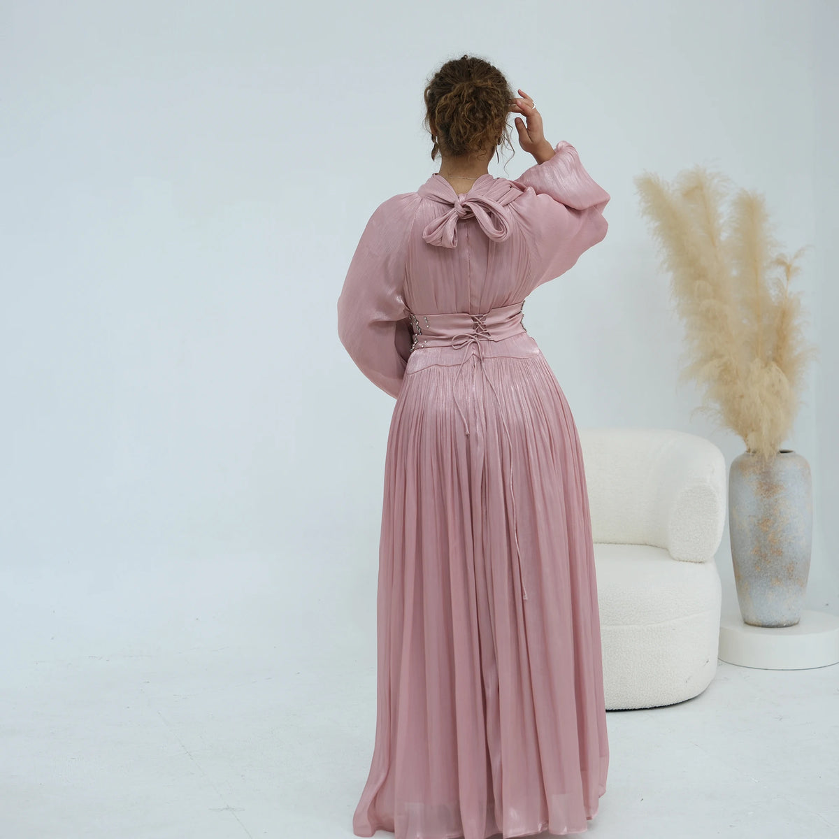 Une femme portant une abaya rose poudré fluide avec une ceinture ornée, debout sur un fond blanc.
