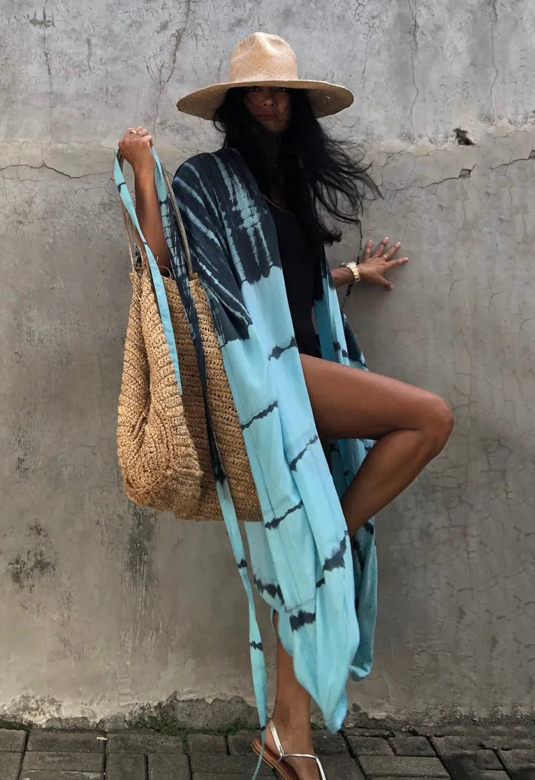 Eine Frau in einem blau-schwarz gemusterten Kimono-Überkleid und einer Strohtasche steht vor einer strukturierten Wand.