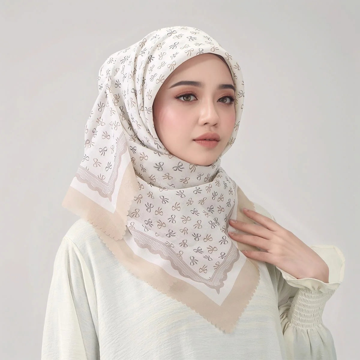 Eine Frau trägt einen hellbeigen floralen quadratischen Hijabschal und ein passendes Oberteil und blickt in die Kamera.