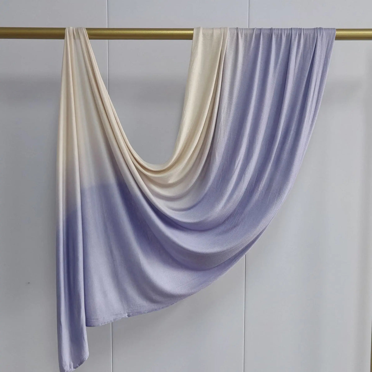 Pañuelo de algodón en degradado lila claro y crema drapeado sobre una varilla dorada.