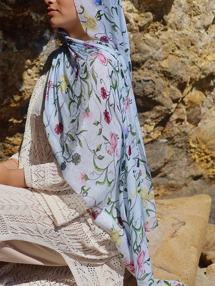 Une femme portant un hijab bleu clair à motifs floraux, assise devant un mur de pierre texturé.
