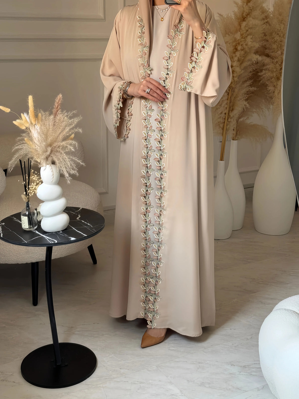 Eine Frau in einem eleganten beige Abaya mit dezentem Blattstickerei steht in einem hellen Innenraum.