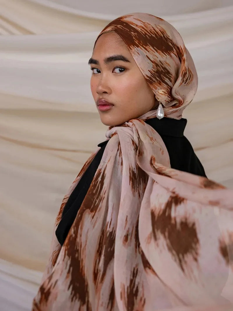 Une femme portant un foulard hijab en polyester doux avec des motifs abstraits marron et beige sur un fond clair.