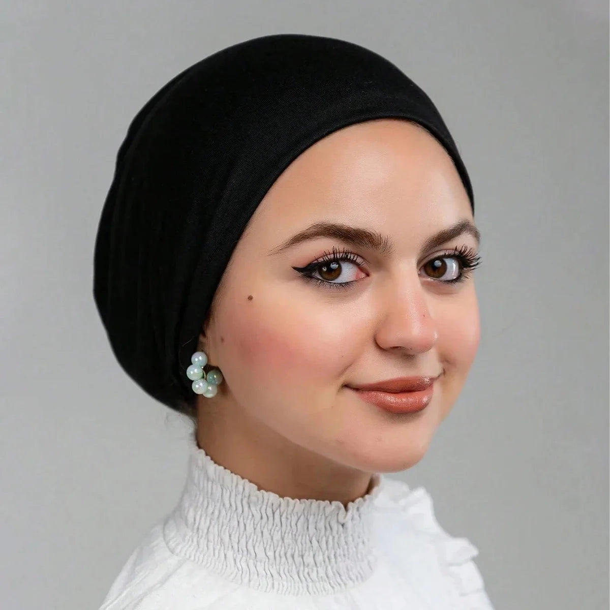 Eine Frau mit schwarzer Stretch-Jersey-Innhijab-Kappe und weißem Oberteil, die in die Kamera blickt.