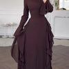 A model wears an elegant, long-sleeved, dark plum A-line maxi dress with ruffled details in a bright room.