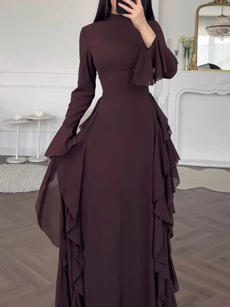 A model wears an elegant, long-sleeved, dark plum A-line maxi dress with ruffled details in a bright room.
