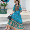 Eine Frau in einem blau-grünen, bestickten Boho-Maxikleid mit Strohhut auf Außentreppen.