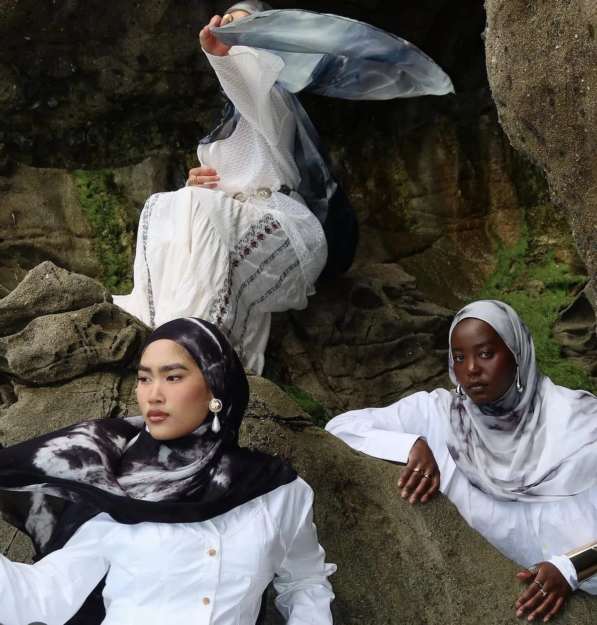 Eine Frau mit weißem Hemd und schwarzem Hijab sitzt auf Felsen, während eine andere Person ein blaues Tuch hält.