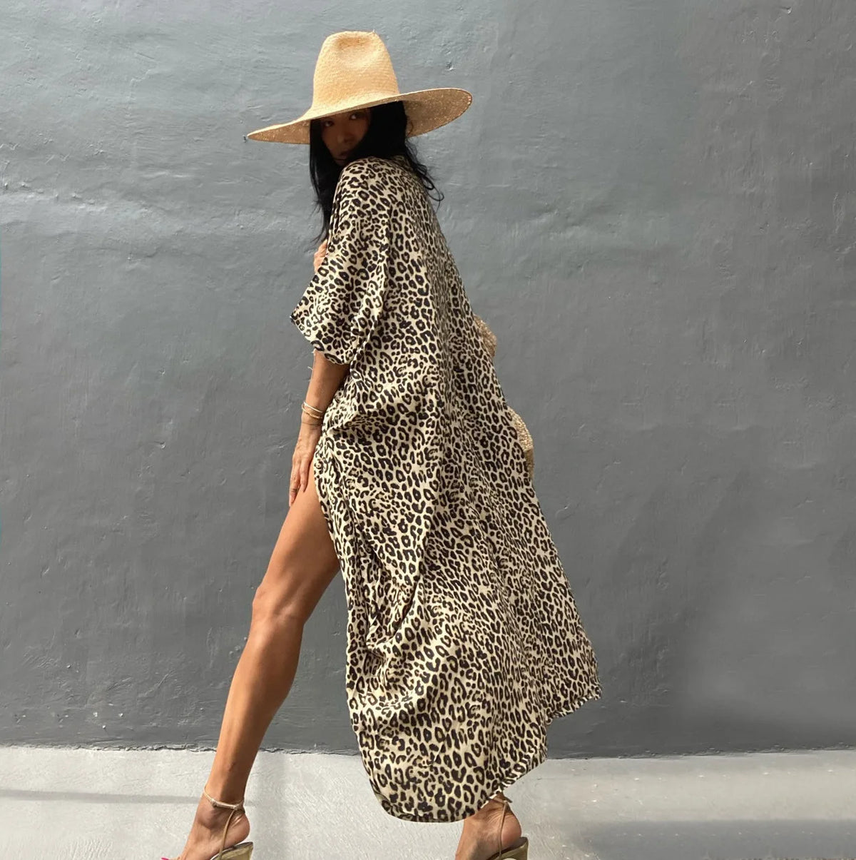 Eine Frau mit Leopardenmuster-Sommer-Strandkimono und Strohhut vor einer grauen Wand.