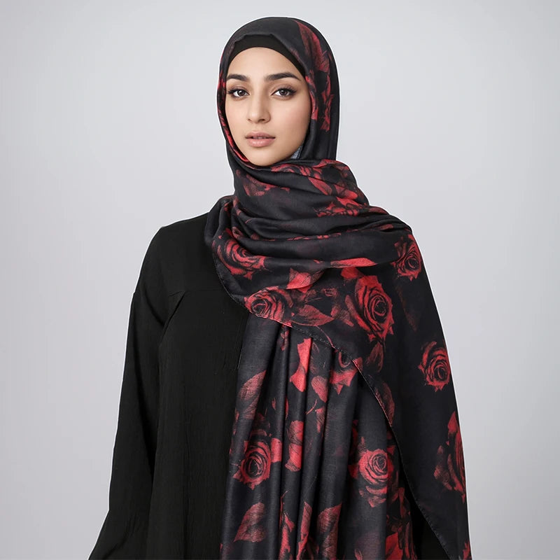 Eine Frau trägt einen schwarzen Hijabschal mit rotem Rosen-Aquarellmuster vor einem hellgrauen Hintergrund.