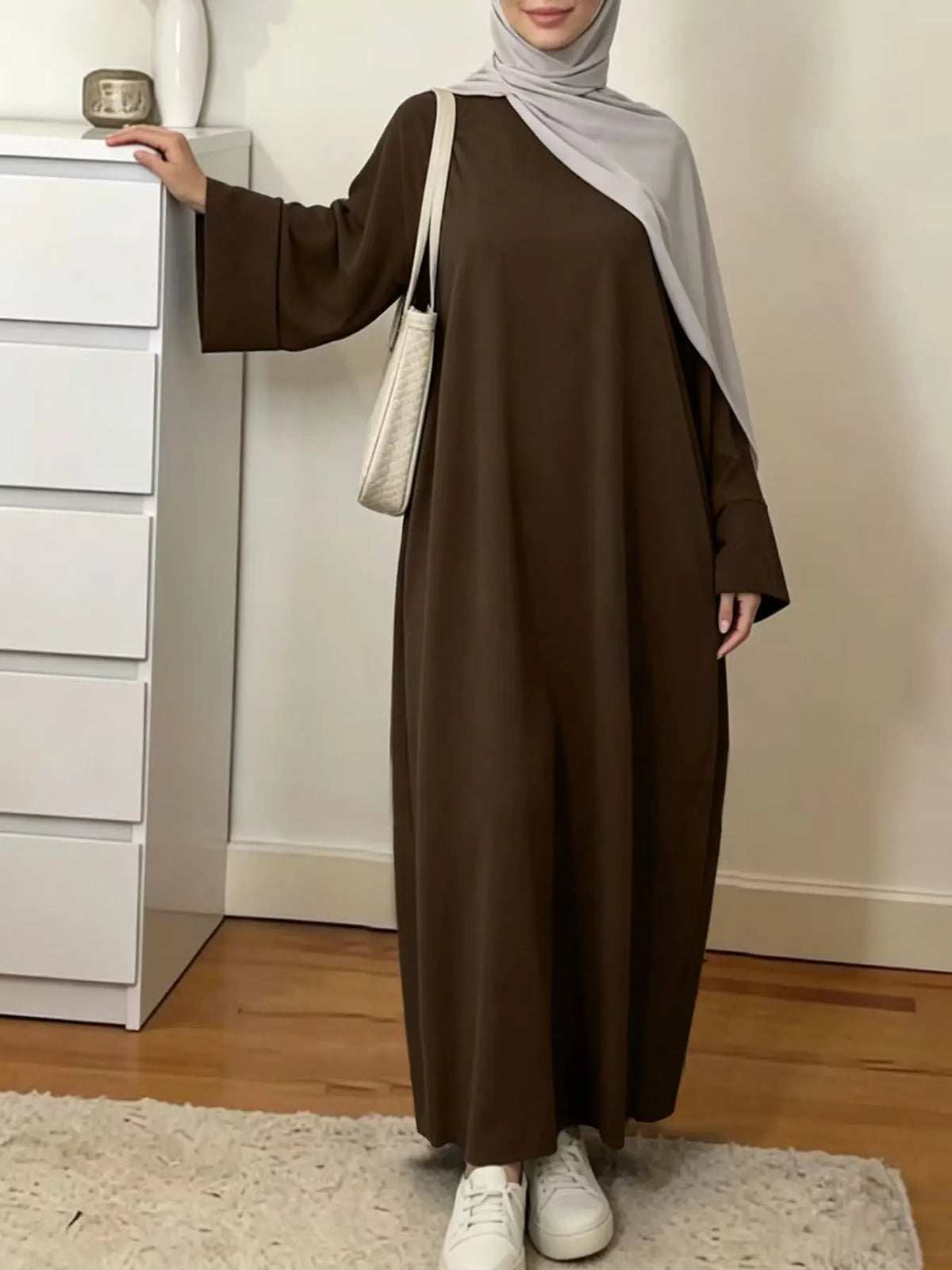 Une femme portant une élégante abaya ample marron uni et un hijab gris clair se tient à côté d'une armoire blanche.