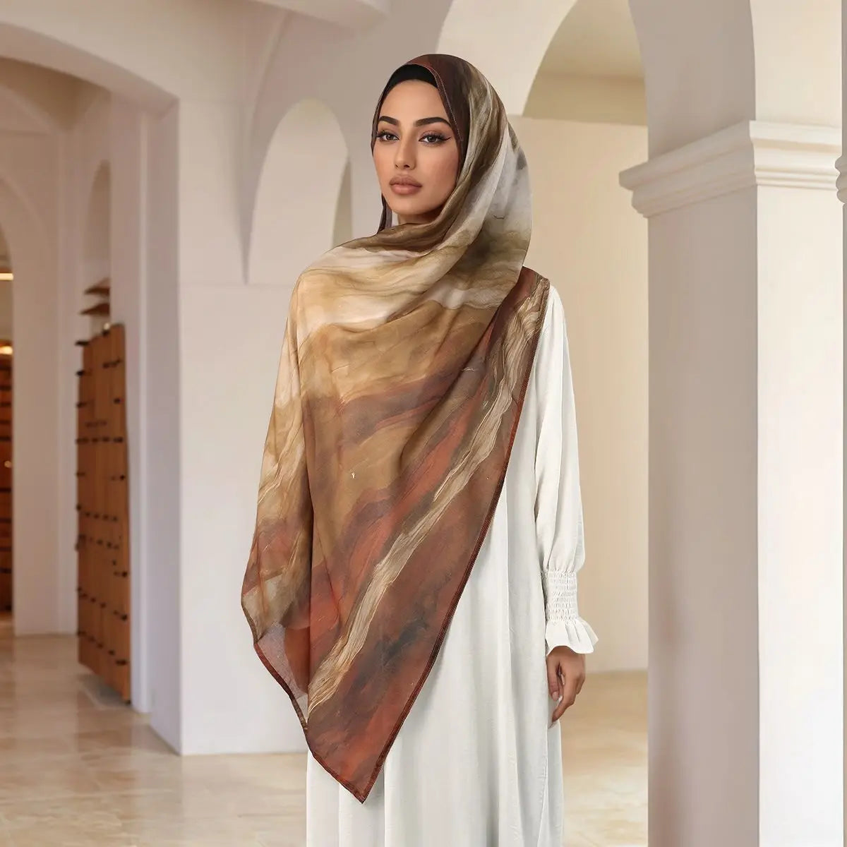 A woman wearing a flowing beige watercolor print modal scarf draped over a white dress in an arched hallway.