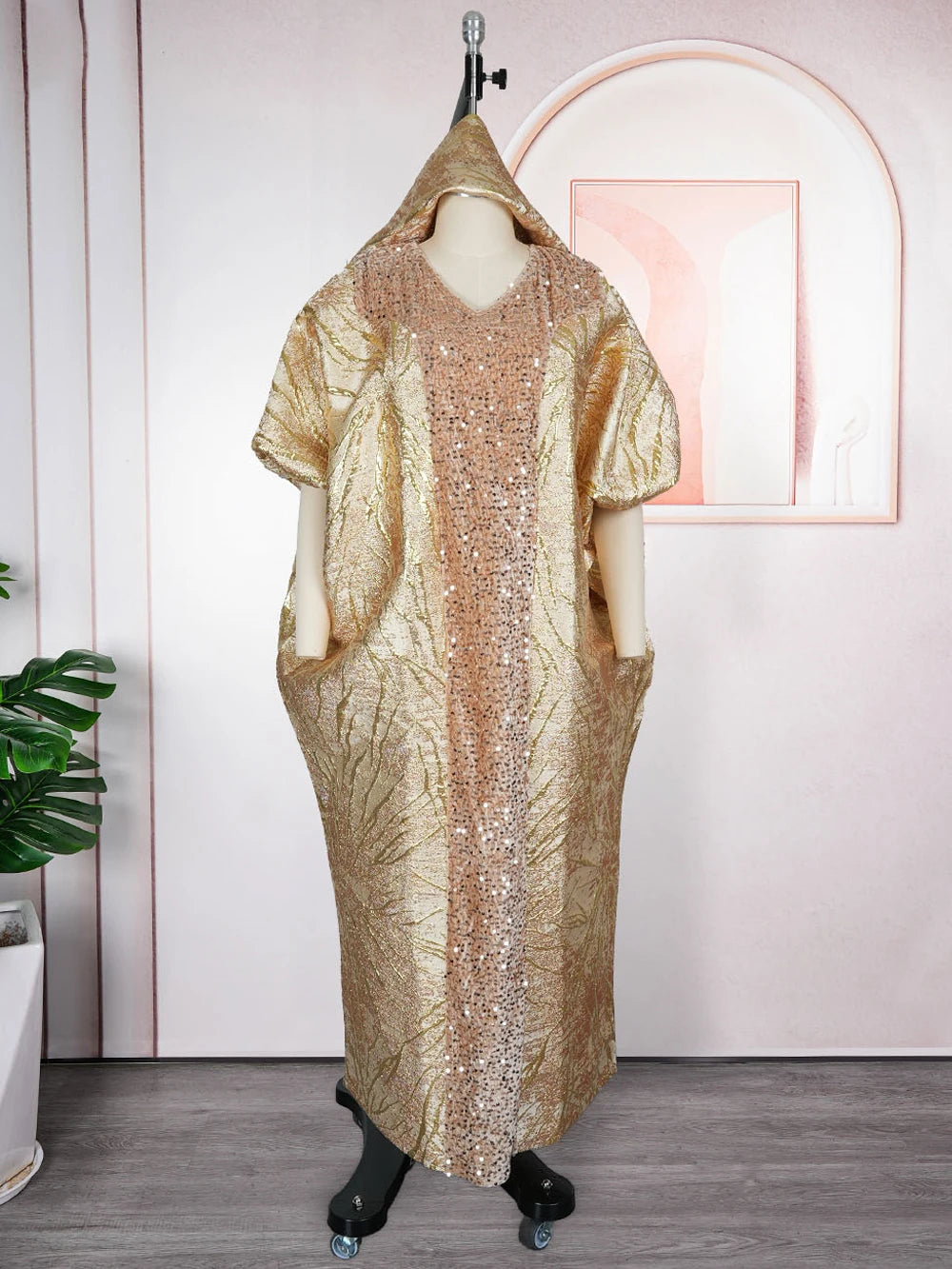 Vestido largo de lentejuelas doradas con capucha, exhibido en maniquí contra una pared rosa.