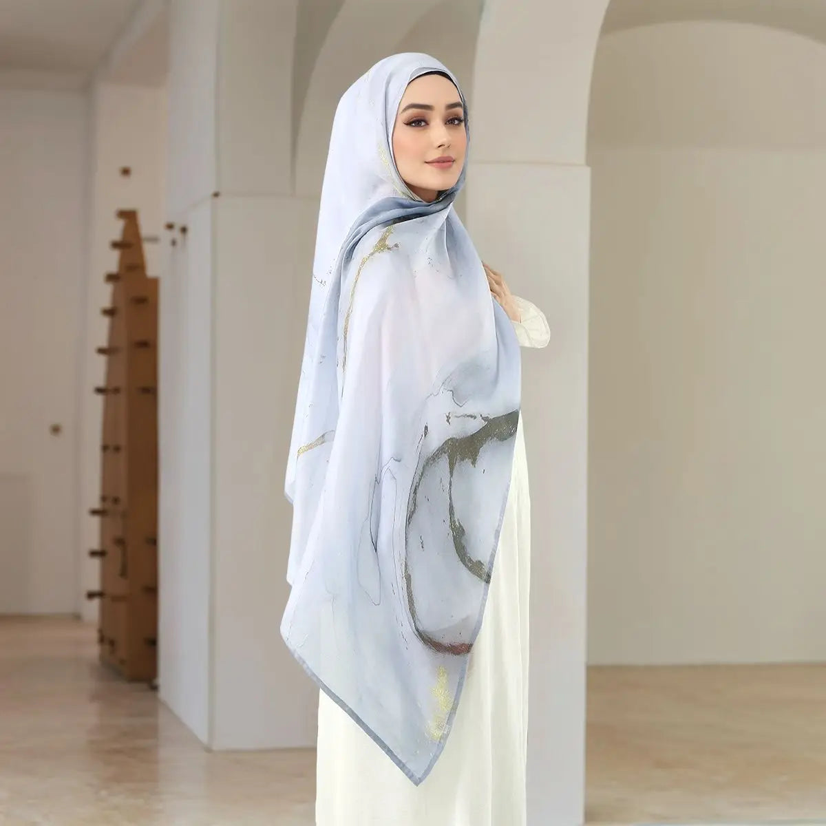Eine Frau trägt einen hellblauen Aquarelldruck-Baumwoll-Leinen-Hijabschal in einem minimalistischen Innenraum.