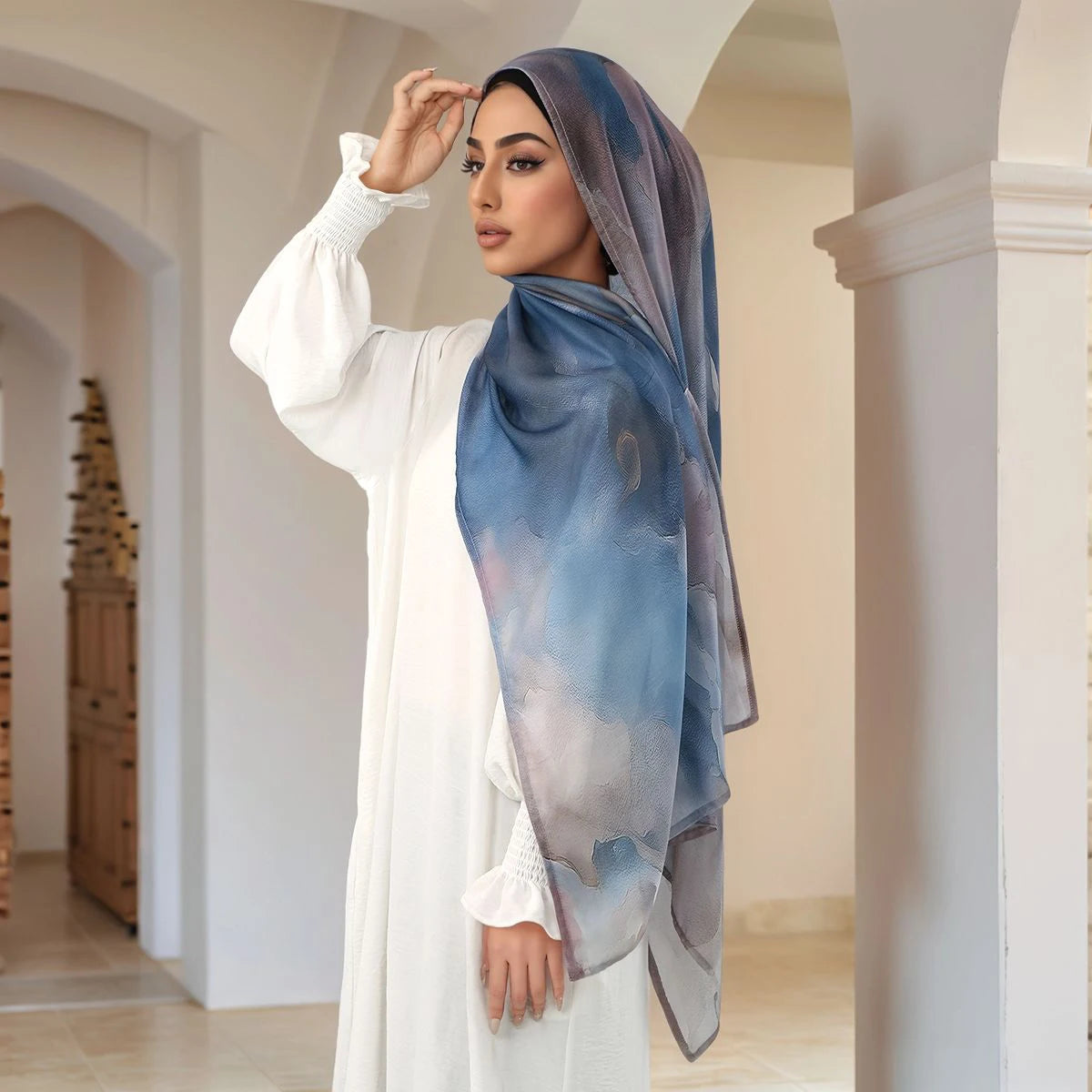 Eine Frau trägt einen blauen Aquarell-Digitaldruck-Hijab und ein weißes traditionelles Kleid in einem gewölbten Flur.