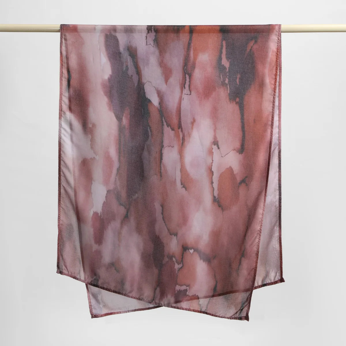 A sheer, printed linen hijab scarf in shades of dusty rose and brown is displayed hanging on a rod.
